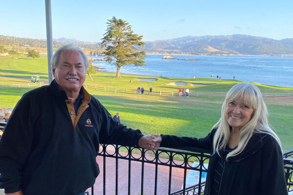 Man and woman standing at railing holding hands, with golf course and ocean in the background