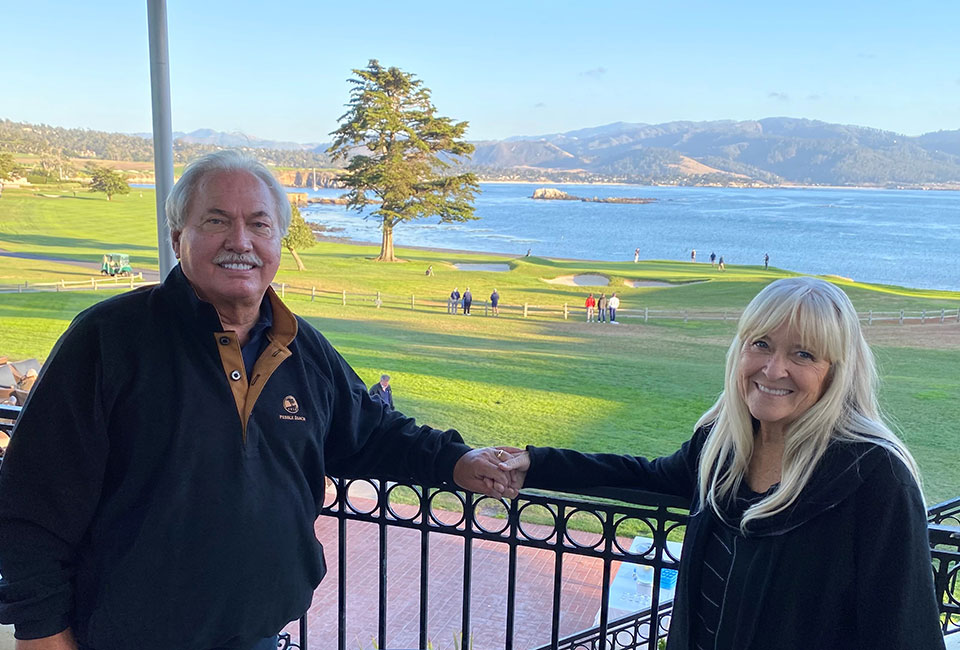 Man and woman standing at railing holding hands, with golf course and ocean in the background